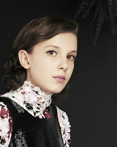 Close-up portrait of a young woman with short brown hair, wearing a black sleeveless dress with a white floral lace collar, against a black background.