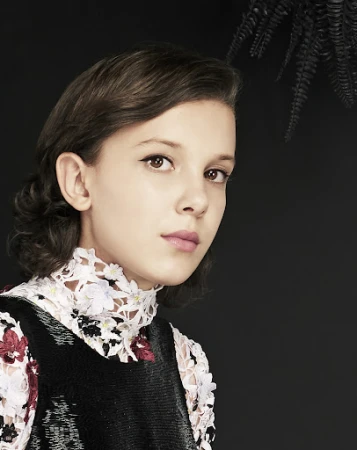 Close-up portrait of a young woman with short brown hair, wearing a black sleeveless dress with a white floral lace collar, against a black background.