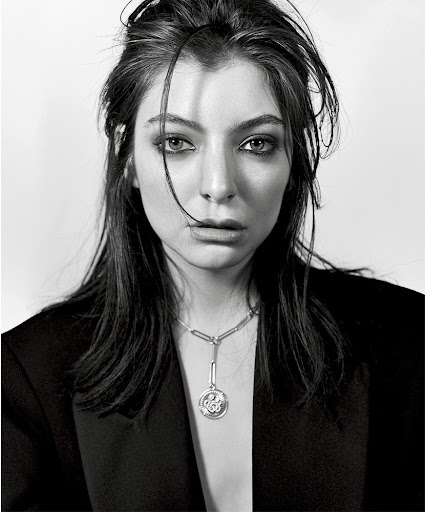 Close-up monochrome portrait of singer Lorde wearing a dark blazer and pendant necklace with smokey eye makeup against a white backdrop