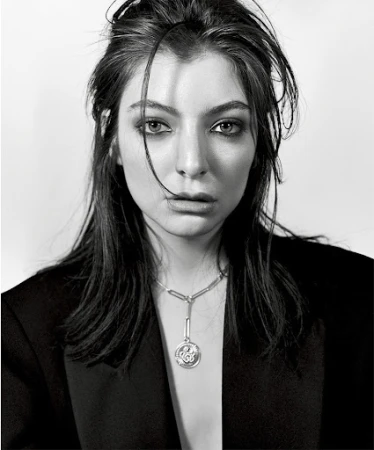 Close-up monochrome portrait of singer Lorde wearing a dark blazer and pendant necklace with smokey eye makeup against a white backdrop