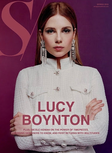 Close-up fashion magazine cover of Lucy Boynton wearing a white boucle jacket and long chandelier earrings against a purple background.