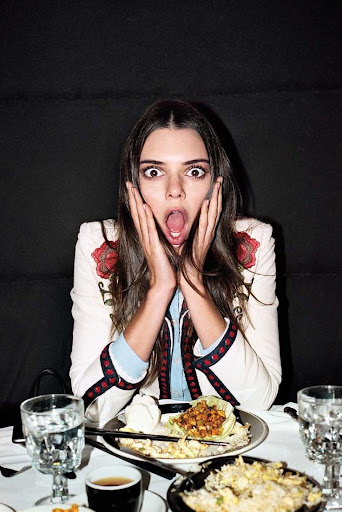 Portrait of a surprised woman with her hands to her face, seated at a dinner table with plates of food and a black background.