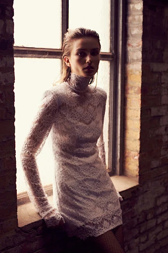 A woman with wet hair poses in a lacy white dress by a window with a brick wall. Fashion shot, neutral expression, and cool tones.