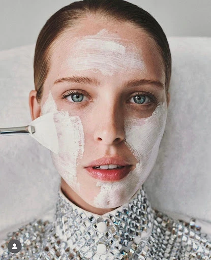 Close-up of a woman receiving a facial treatment with a white cream mask applied by a brush in a bright, indoor setting.