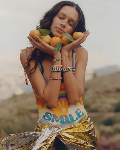 Woman poses with lemons outdoors, wearing beaded bracelets and a dress with 'SMILE' on it, against a hazy, natural backdrop.