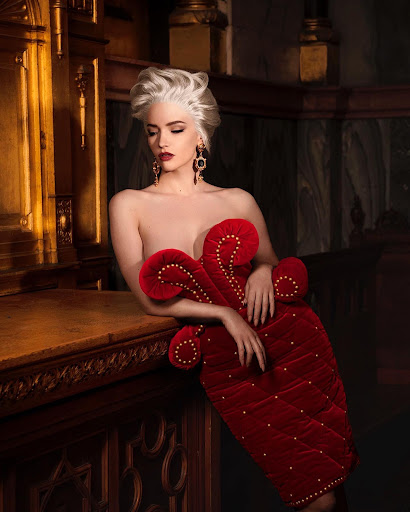 Artistic studio shot of a model with elaborate blonde hair, red lipstick, dramatic red dress, posing against a dark background.