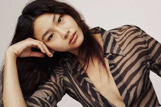 Portrait of an Asian woman wearing a zebra-striped blouse, leaning on her hand with a neutral facial expression, against a plain backdrop.