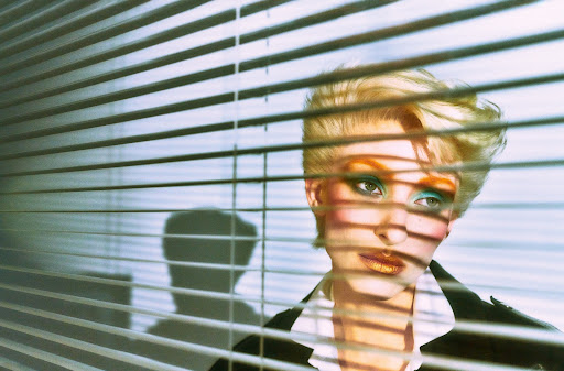 Striking portrait of a stylish woman with short blonde hair, vibrant makeup, and a black jacket, partially obscured by window blinds casting shadows.