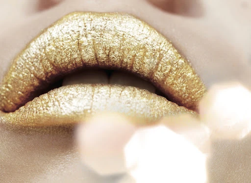 Close-up shot of a woman's lips covered in metallic gold lipstick with bokeh light effects. Focus on the texture and color of the lips.