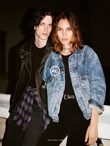 A young man in a black leather jacket and a woman in an oversized blue denim jacket standing close outdoors.