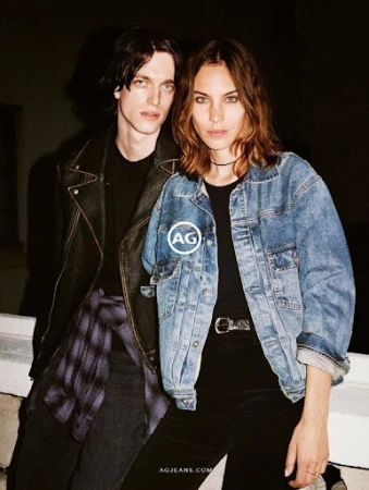 A young man in a black leather jacket and a woman in an oversized blue denim jacket standing close outdoors.
