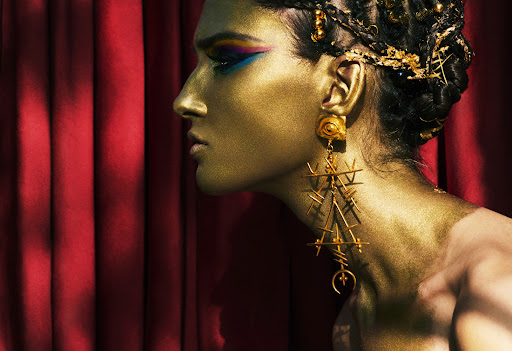 Profile of adult female with face and body painted gold, wearing long gold earrings, styled hair and makeup with a dark red backdrop.