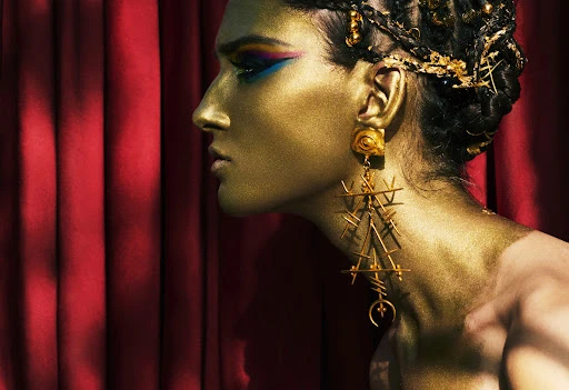 Profile of adult female with face and body painted gold, wearing long gold earrings, styled hair and makeup with a dark red backdrop.