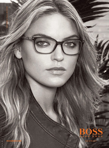 Monochrome portrait of a blonde woman wearing square-framed glasses, a dark jacket, and a neutral expression, in a Boss eyewear ad.