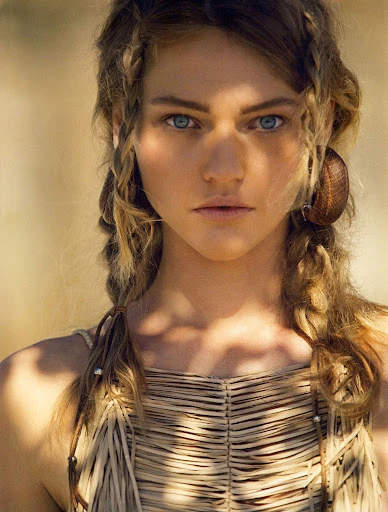 Close-up portrait of a young woman with blue eyes and blonde braids, wearing a woven top, with a blurred outdoor background.