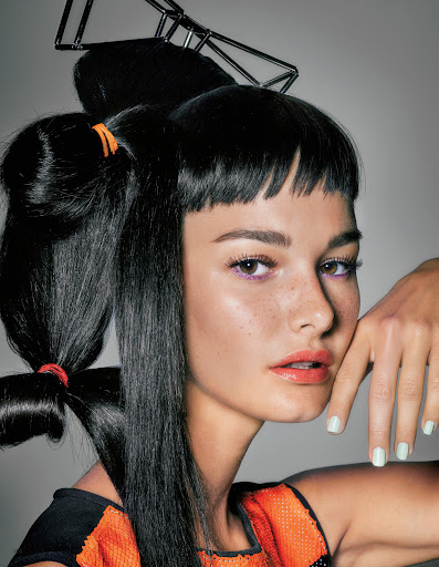 Close-up of a woman with creative black hair styling and an orange beaded top posing against a light gray background.