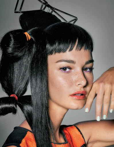 Close-up of a woman with creative black hair styling and an orange beaded top posing against a light gray background.