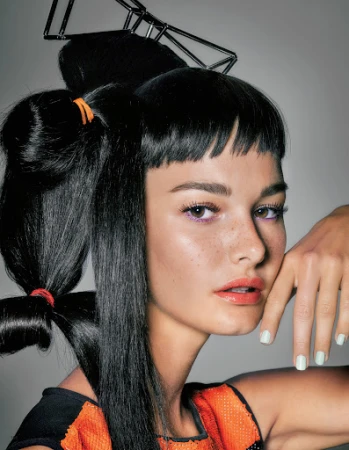 Close-up of a woman with creative black hair styling and an orange beaded top posing against a light gray background.