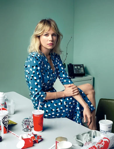 Woman in polka dot dress sits on a desk surrounded by fast food containers in a retro office setting. Medium shot with soft lighting.