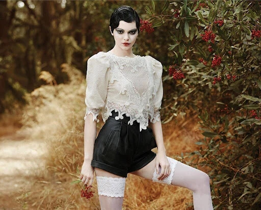Woman in vintage-style fashion with white lace top, black shorts, and thigh-high stockings posing in a forest setting.
