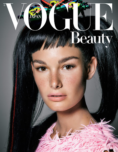 Close-up portrait of a young woman with freckles and black pigtails wearing a pink feathered top against a grey backdrop, Vogue Beauty cover.