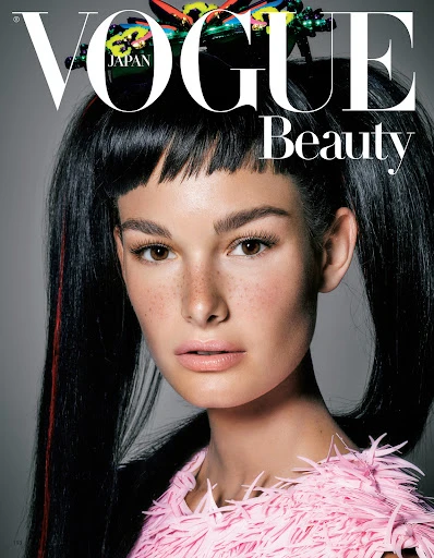 Close-up portrait of a young woman with freckles and black pigtails wearing a pink feathered top against a grey backdrop, Vogue Beauty cover.