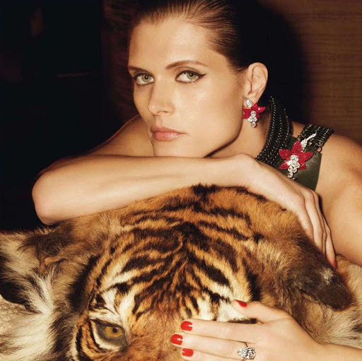 Adult woman with fair skin and light eyes poses with a tiger fur, wearing flower earrings and necklace, against a dark studio background.