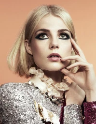 Close-up portrait of a blonde woman with dark smoky eye makeup wearing a sequined garment and ruffled collar against peach background