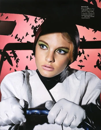 Model with green eyeshadow wearing white leather gloves holding a steering wheel against a pink sky background filled with black silhouettes of birds