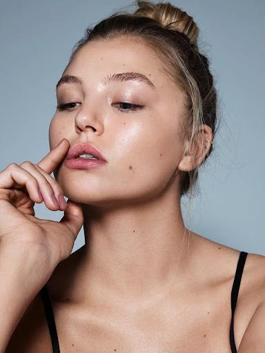 Close-up beauty portrait of a woman with dewy skin and natural makeup touching her lip against a soft grey background.