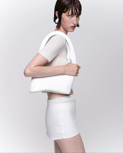 Fashion model in a photo studio wears a beige top, white skirt, white earrings, and carries a white handbag against a white backdrop.