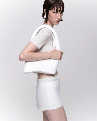 Fashion model in a photo studio wears a beige top, white skirt, white earrings, and carries a white handbag against a white backdrop.