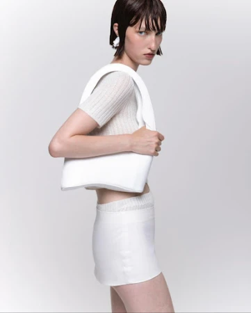 Fashion model in a photo studio wears a beige top, white skirt, white earrings, and carries a white handbag against a white backdrop.