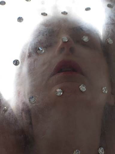 A woman's face is seen looking up through a veil with rhinestones, with a blurred and dreamy aesthetic, against a bright background.