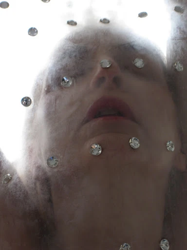A woman's face is seen looking up through a veil with rhinestones, with a blurred and dreamy aesthetic, against a bright background.