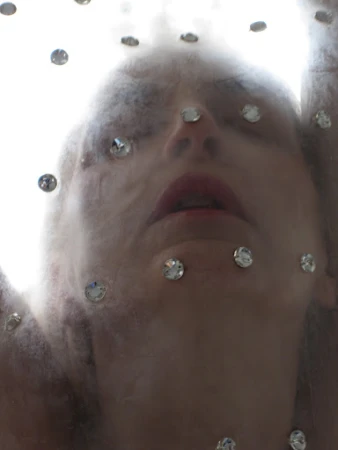 A woman's face is seen looking up through a veil with rhinestones, with a blurred and dreamy aesthetic, against a bright background.