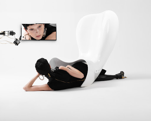 Woman in black outfit lounges by a white chair, watching a woman on TV; robotic arm is connected to the screen, white studio background.