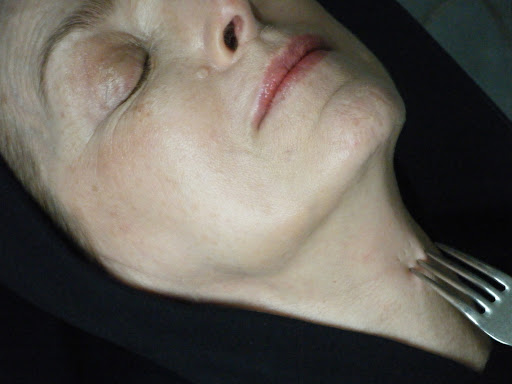 Close-up of a woman's face and neck being touched by a fork. The woman has fair skin, light pink lips, and one eye closed.