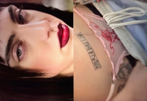 Close-up of a fair-skinned young adult female with bright red lipstick, and pink lingerie with a tattoo, against a soft-focus background.
