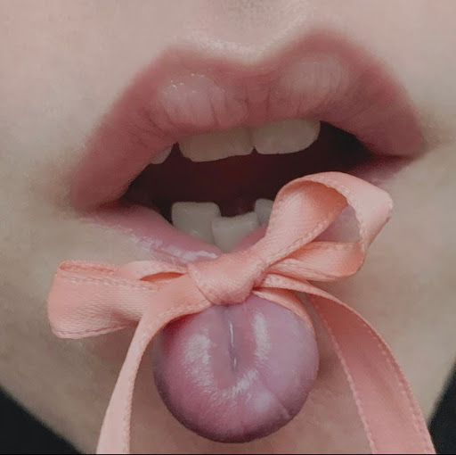 Close-up of a person's mouth with open lips, visible teeth, and tongue playfully adorned with a tied light pink bow.