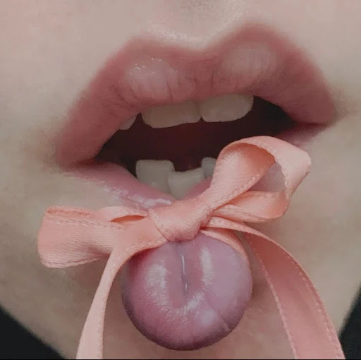 Close-up of a person's mouth with open lips, visible teeth, and tongue playfully adorned with a tied light pink bow.