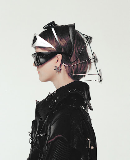 Side profile portrait of a person wearing a black jacket, star-shaped earrings, and avant-garde headwear against a white background.