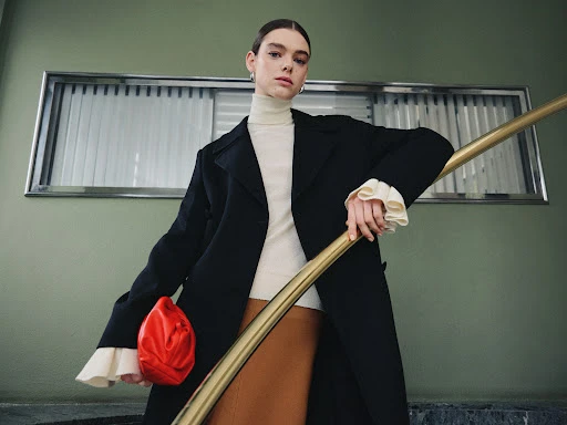 A stylish woman in a black coat, beige turtleneck, and brown skirt poses on a staircase with a bright red clutch.