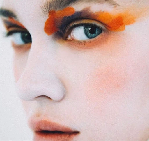 Close-up portrait of a young adult female with striking blue eyes and artistic orange and purple eyeshadow, against a light backdrop.