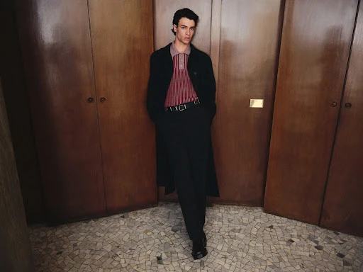 Fashionable young adult male standing indoors wearing a long coat, vertically striped polo shirt, black dress pants, and black dress shoes.