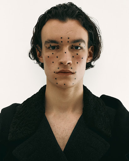 Portrait of a young adult man with black hair and dotted makeup, wearing a dark coat, against a plain white backdrop.