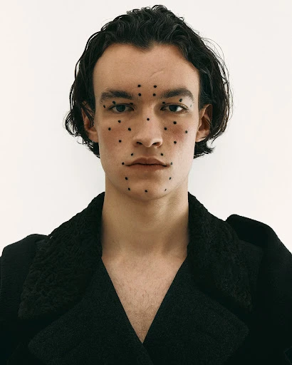 Portrait of a young adult man with black hair and dotted makeup, wearing a dark coat, against a plain white backdrop.