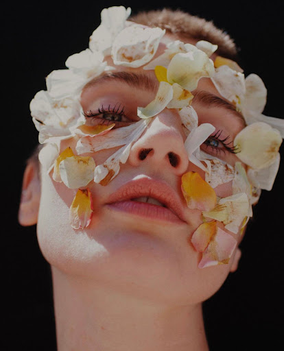 Close-up of a woman's face partially covered in white and yellow flower petals against a dark black background in soft, diffused lighting.