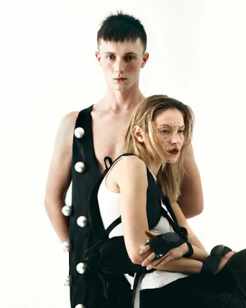 A young male model with dark hair wearing a black garment with large pearl embellishments standing behind a female model with facial dots and dark lipstick.
