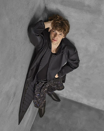 Eye-level shot of a light-skinned male model with curly hair in a black jacket and printed pants leaning against a grey corner.